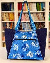 Load image into Gallery viewer, Blue floral tote bag with navy accents on a white surface in front of a bookshelf.