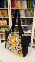 Load image into Gallery viewer, Floral tote bag with black accents on a white surface in front of a bookshelf.
