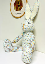 Load image into Gallery viewer, Stuffed bunny toy with geometric pattern on a white surface, with a patchwork wall hanging in the background.
